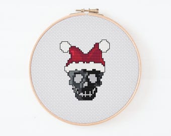 May include: Cross-stitch embroidery of a skull wearing a Santa hat, displayed in a wooden hoop. The skull is black and gray, and the hat is red and white. The background is a light gray fabric.