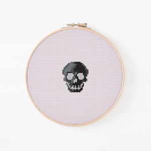 May include: A cross-stitch embroidery of a skull in a wooden hoop. The skull is black and gray, with white eye sockets and teeth. The background is a light pink fabric.
