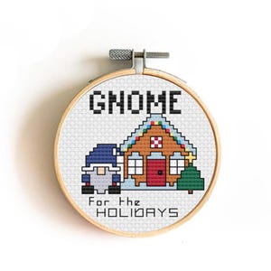 May include: Cross-stitch embroidery in a wooden hoop features a pixelated gnome and house design. The text "GNOME" is above the house, and "For the HOLIDAYS" is below. The gnome wears a blue hat and has a gray beard. The house has a red door and a green Christmas tree.