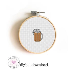 Op de afbeelding: Een borduurring met kruissteek met een gepixeld bierpulontwerp. De bierpul is bruin met een grijze schuimtop. De tekst "digital download" staat onder een roze hartlogo. De houten ring is vastgezet met een metalen sluiting.
