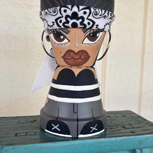 May include: A hand-painted terracotta pot shaped like a figure wearing a black and white bandana, a black tube top with white stripes, and grey pants. The figure features large brown eyes, dark lips, and silver hoop earrings attached to the sides.