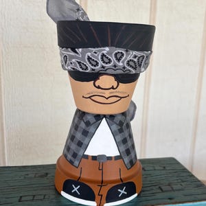 May include: A hand-painted terracotta pot designed to resemble a figure wearing a grey paisley bandana, black sunglasses, a grey and black checkered flannel shirt over a white t-shirt, brown pants, and black sneakers with white X details.