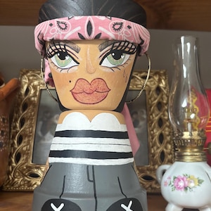 May include: A hand-painted terracotta pot shaped like a person with dark hair, a pink bandana, and large gold hoop earrings. The figure features green eyes, full lips, a white top, and grey pants with black shoes marked with white X symbols.
