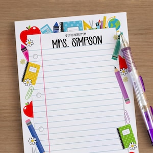 May include: A notepad with blue lined paper and a decorative border of school supplies, including apples, pencils, and notebooks. The text "A little note from Mrs. Simpson" is printed at the top. A purple pen rests on the notepad.