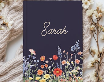 May include: A dark purple hardcover journal featuring the name Sarah in elegant gold script lettering. The bottom portion of the cover is decorated with a colorful botanical illustration of various wildflowers in shades of red, orange, yellow, and blue.