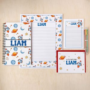 May include: A stationery set featuring a space theme with rockets, planets, and stars. The set includes a spiral notebook, lined notepad, sticky notes, and a greeting card, all personalized with the name "LIAM" in blue. A multi-colored pen is also included.