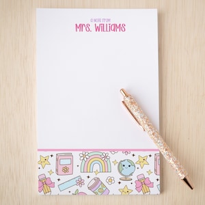 May include: A white notepad with the text A NOTE FROM MRS. WILLIAMS in pink at the top and a colorful border at the bottom featuring illustrations of rainbows, books, globes, and pencils, accompanied by a rose gold glittery ballpoint pen.