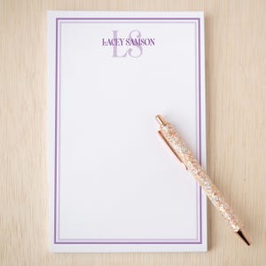 May include: White notepad with a purple border and the text "LACEY SAMSON" and initials "LS" in purple. A rose gold glitter pen rests on the notepad. The notepad and pen are on a light wood surface.