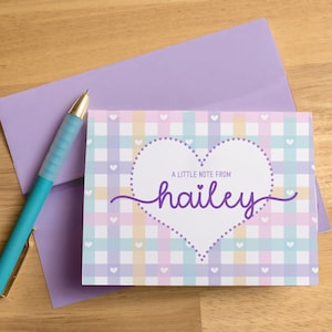 May include: A pastel-colored stationery set featuring a greeting card and envelope. The card has a gingham pattern with a heart-shaped design and the name "hailey" in cursive. A teal pen rests beside the stationery.