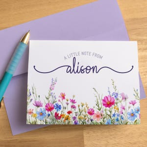 May include: A white stationery card with a floral design and the text "A LITTLE NOTE FROM alison". The card is decorated with colorful flowers in pink, blue, and yellow. A light purple envelope and a teal pen are also visible.