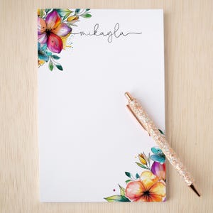 May include: White notepad with floral watercolor design in the corners, featuring orange, pink, and blue flowers with green leaves. The name "mikayla" is written in cursive. A rose gold glitter pen rests on the notepad.