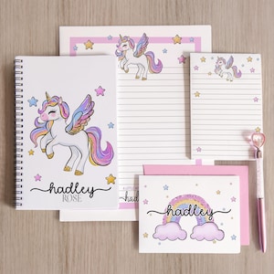 Peut inclure: Ensemble de papeterie sur le thème de la licorne. Il comprend un carnet à spirales, du papier ligné, des blocs-notes, une carte de vœux et un stylo. Les articles sont blancs avec des illustrations de licornes, des étoiles et le nom "Hadley Rose". Le stylo a un cœur.