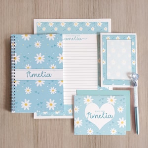 May include: A stationery set with a light blue daisy design. The set includes a spiral-bound notebook, lined paper, a notepad, notecards, and a pen. The name "Amelia" is printed on the notebook, notecards, and lined paper.