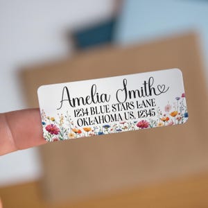 May include: A rectangular white label with floral border and the name "Amelia Smith" in elegant script. The label includes an address: 1234 Blue Stars Lane, Oklahoma US, 12345. The floral border features colorful wildflowers.