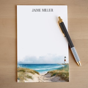 May include: A white notepad with the name "JAIME MILLER" printed at the top. A watercolor painting of a beach scene with a lighthouse is at the bottom. A black pen with gold accents rests on the notepad.