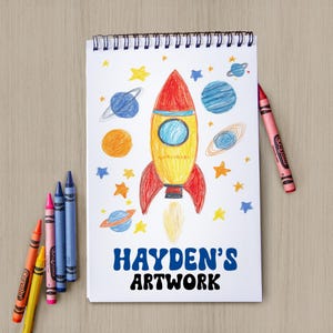 May include: A spiral-bound sketchbook featuring a hand-drawn illustration of a yellow and red rocket ship surrounded by blue, orange, and yellow planets and stars. The text "HAYDEN'S ARTWORK" is printed in bold blue and black letters at the bottom.
