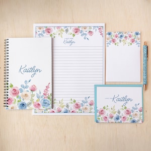 May include: A stationery set featuring a notebook, notepad, lined paper, and a card, all adorned with watercolor floral designs in pink, blue, and white. The name "Kaitlyn" is written on each item. A matching pen is also included.