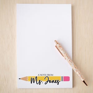 May include: A rectangular white notepad featuring a graphic of a yellow pencil with a pink eraser at the bottom. The text "A NOTE FROM" is printed above the pencil, and "Ms. Jones" is written in black script across the pencil. A rose gold glitter pen rests on the notepad.