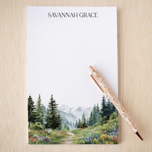 May include: A notepad with the text "SAVANNAH GRACE" at the top. The bottom of the notepad features a watercolor painting of a mountain landscape with trees and a path. A glittery pen rests on the notepad.
