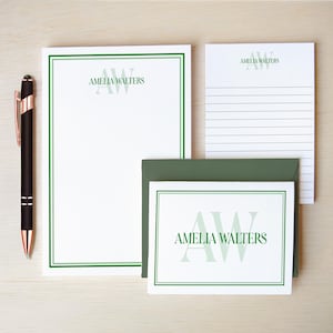 May include: A stationery set featuring a large white notepad, a smaller lined notepad, and a note card with a dark green envelope. Each piece displays the name AMELIA WALTERS and the initials AW in green. A black pen with rose gold accents is included.