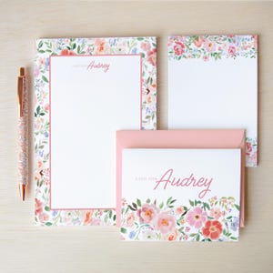 May include: A stationery set featuring a notepad, a smaller memo pad, and a folded note card with a pink envelope, all decorated with watercolor floral borders. The notepad reads A NOTE FROM Aubrey and the note card reads A NOTE FROM Audrey, with a glittery pen.