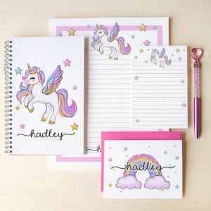 May include: A stationery set featuring a spiral-bound notebook, a lined notepad, and a greeting card, all decorated with white unicorns with rainbow manes and wings, colorful stars, and the name hadley. A pink heart-topped pen is included.