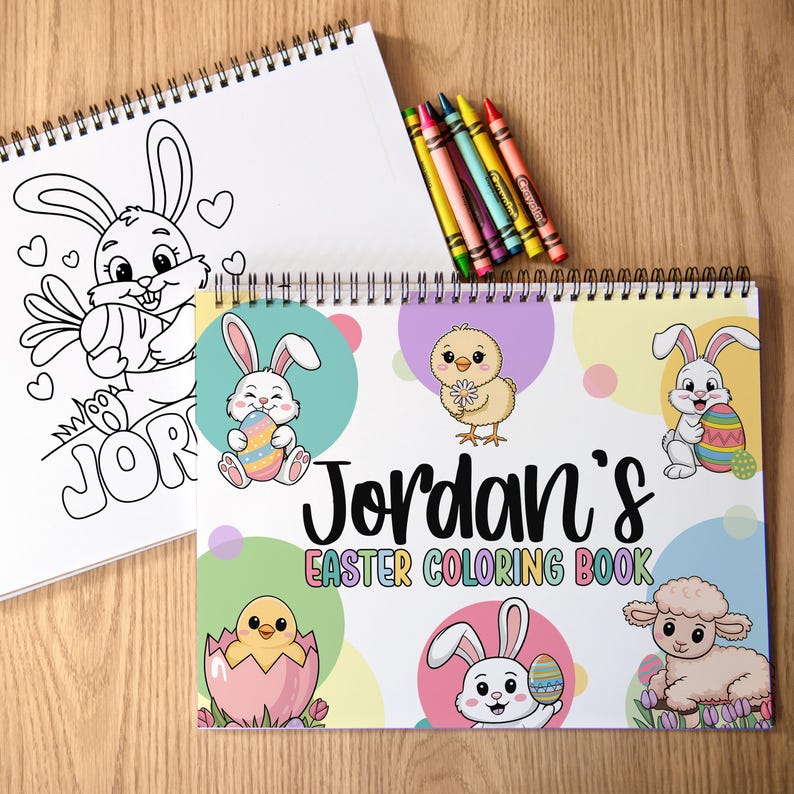 K&ouml;nnte beinhalten: Zwei Spiral-Malb&uuml;cher und Buntstifte auf einer Holzoberfl&auml;che. Ein Buch zeigt eine Schwarz-Wei&szlig;-Hasenillustration mit dem Namen "Jordan". Das andere Buch tr&auml;gt den Titel "Jordan's Easter Coloring Book" und enth&auml;lt farbenfrohe Illustrationen zum Thema Ostern.