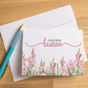 May include: A white stationery set with a floral design and the text "A note from Lauren." The set includes a card, envelope, and a teal pen with gold accents. The card features pink gladiolus flowers.