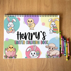 K&ouml;nnte beinhalten: Ein Spiral-Malbuch mit dem Text "Henry's Easter Coloring Book" und Illustrationen von Osterhasen, K&uuml;ken und einem Lamm. Buntstifte in verschiedenen Farben liegen auf der Holzoberfl&auml;che neben dem Buch.