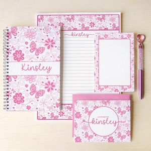 May include: A stationery set featuring a spiral-bound notebook, a lined notepad, a small notepad, and a greeting card, all decorated with a pink butterfly and floral pattern. The name Kinsley is printed on the notebook, notepad, and card.