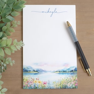 May include: A white notepad with the name "mikayla" in blue script. The bottom of the notepad features a watercolor landscape of a lake, mountains, and colorful flowers. A black and gold pen rests on the notepad.