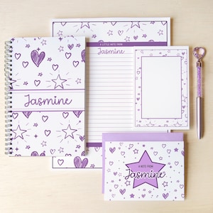 May include: A stationery set featuring a spiral-bound notebook, a lined notepad, a small notepad, and a greeting card with a purple heart and star pattern. Each item is personalized with the name Jasmine. A purple pen with a heart topper is included.