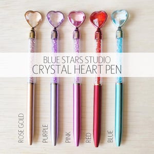 A collection of five colorful heart-shaped pens with glittery centers, arranged on a wooden surface.