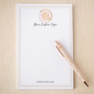 May include: A white rectangular notepad with a thin gold border, featuring a gold floral logo in a pink circle, the text "Your Custom Logo", and the name "AMANDA WILLIAMS" at the bottom, accompanied by a rose gold glitter ballpoint pen.