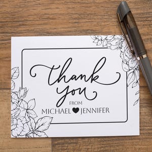 May include: A white card with a black border and floral corner designs. The card features the words "Thank you" in elegant script, with "from MICHAEL ♥ JENNIFER" below. A gray pen rests on the wooden surface.