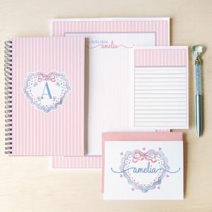 May include: A stationery set featuring a spiral-bound notebook, a notepad, a letterhead, and a greeting card, all with pink and white vertical stripes. The items display a heart-shaped lace graphic with the letter A or the name amelia, plus a pen.