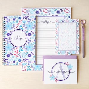 May include: A stationery set featuring a spiral-bound notebook, a lined notepad with the text A LITTLE NOTE FROM ashlyn, a small notepad, a greeting card with the name ashlyn and letter A, and a pen with a heart-shaped topper, all with floral patterns.