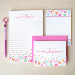 May include: A stationery set including a lined notepad, a blank notepad, and a note card with a matching pink envelope. Each item features a colorful heart pattern and the text A LITTLE NOTE FROM madison. A pink pen with a heart topper is included.