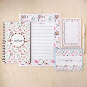 May include: A stationery set featuring a spiral-bound notebook, a lined notepad with the text A NOTE FROM heather, a small notepad, and a folded card with the text A NOTE FROM heather, all decorated with a floral pattern of pink and purple flowers.