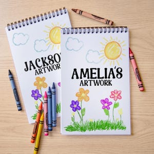 May include: Two spiral-bound art notebooks with the text "JACKSON'S ARTWORK" and "AMELIA'S ARTWORK". Each notebook features a sun, clouds, and flower drawings. Crayons in various colors are scattered around the notebooks.