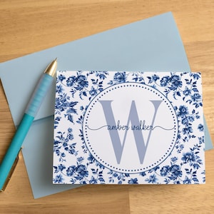 May include: A rectangular note card featuring a blue and white floral pattern with a central dotted circle containing a large letter W and the name amber walker in script. A light blue envelope and a turquoise pen are positioned next to the card.