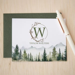 May include: A white stationery card featuring a green letter W inside a circular branch frame, the name Taylor Walters, and a watercolor mountain and pine forest landscape at the bottom, paired with a dark green envelope and a white pen.