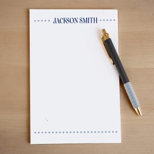 May include: A white notepad with the name "JACKSON SMITH" in navy blue, dotted borders, and a black pen with gold accents. The pen rests on the notepad, which is on a light wood surface.