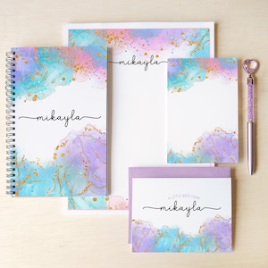 May include: A stationery set featuring a spiral-bound notebook, a notepad, a letterhead, and a note card with envelopes, all decorated with a teal, pink, and purple watercolor wash and gold glitter accents. Each item displays the name mikayla.