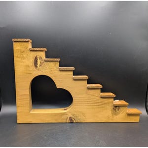 May include: A wooden staircase display stand featuring a natural light brown finish with visible wood grain and knots. The structure includes seven tiered steps and a large, heart-shaped cutout opening in the center of the main side panel.