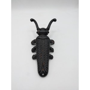 May include: A black cast iron boot jack shaped like a beetle. The insect-shaped tool features two long, curved antennae at the top, a textured wing casing with a repeating scale pattern, and six small legs along the sides of the elongated body.