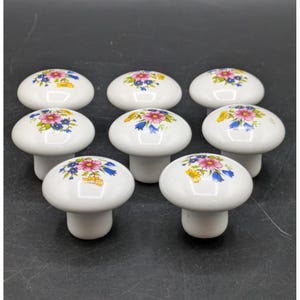 May include: A set of seven white ceramic cabinet knobs, each featuring a rounded top decorated with a colorful floral pattern of pink, blue, and yellow flowers with green leaves. The knobs have a smooth, glossy finish and a cylindrical base.