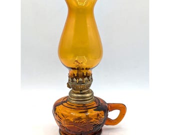 Puede incluir: Una lámpara de aceite de vidrio ámbar con una base texturizada y estampada, un asa lateral y un quemador metálico. La lámpara está rematada con una chimenea de vidrio ámbar alta y lisa que se ensancha ligeramente en la abertura superior.