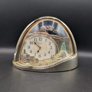 May include: A gold-toned Rhythm Quartz clock with a clear, arched top. The clock face is white with black numerals and hands. The base features a detailed cityscape and a small statue. The clock is on a black surface.