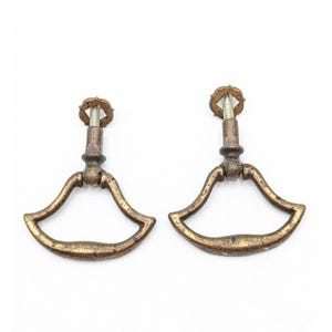 May include: Two brass-toned metal drawer pulls with a fan-shaped, curved handle design. Each pull features a central vertical post topped with a small, decorative circular nut and a threaded metal screw extending from the back for mounting purposes.